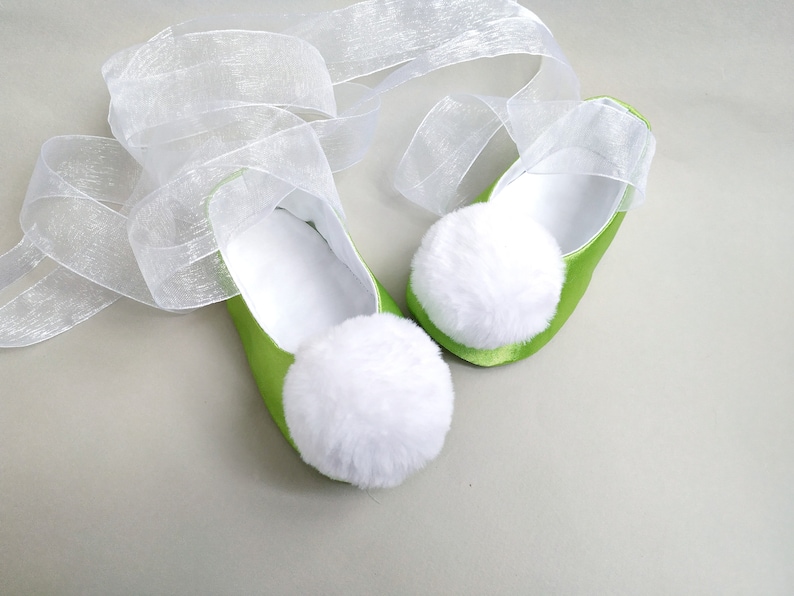May include: A pair of green satin baby shoes with white pom-poms and a white tulle ribbon.