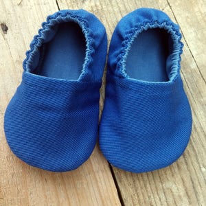Op de afbeelding: Een paar blauwe babyschoentjes van zachte stof. De schoentjes hebben een ronde neus en elastiek rond de opening. Ze worden getoond op een houten oppervlak.