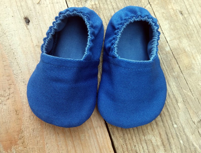 Op de afbeelding: Een paar blauwe babyschoentjes van zachte stof. De schoentjes hebben een ronde neus en elastiek rond de opening. Ze worden getoond op een houten oppervlak.