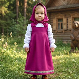 Può includere: Una bambina indossa un vestito magenta, una camicetta bianca e un cappuccio abbinato. L'abito presenta un bordo decorativo sull'orlo. La bambina è all'aperto, con un edificio in legno sullo sfondo.