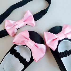 Peut inclure: Un ensemble de chaussures de b&eacute;b&eacute; noires avec des n&oelig;uds en satin rose. Les chaussures ont des bandes &eacute;lastiques autour des chevilles. Un bandeau noir avec un n&oelig;ud en satin rose est &eacute;galement inclus.