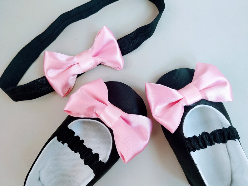 Peut inclure: Un ensemble de chaussures de b&eacute;b&eacute; noires avec des n&oelig;uds en satin rose. Les chaussures ont des bandes &eacute;lastiques autour des chevilles. Un bandeau noir avec un n&oelig;ud en satin rose est &eacute;galement inclus.