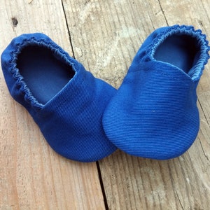 Op de afbeelding: Een paar blauwe babyschoentjes van een zachte, getextureerde stof. De schoentjes hebben een ronde neus en een elastische opening. De schoentjes zijn ontworpen voor comfort en zijn waarschijnlijk bedoeld voor gebruik binnenshuis. De schoentjes liggen op een houten oppervlak.