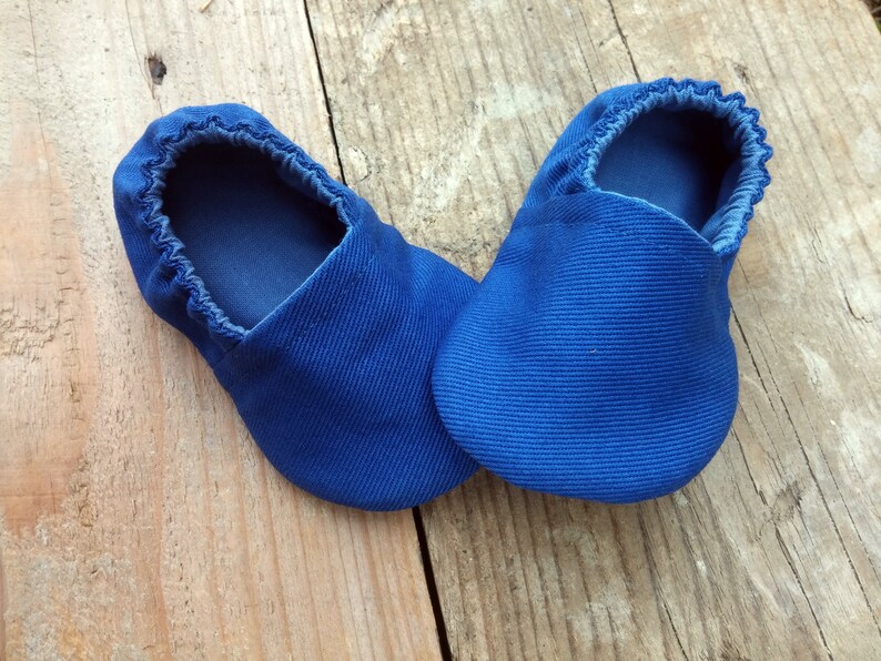 Op de afbeelding: Een paar blauwe babyschoentjes van een zachte, getextureerde stof. De schoentjes hebben een ronde neus en een elastische opening. De schoentjes zijn ontworpen voor comfort en zijn waarschijnlijk bedoeld voor gebruik binnenshuis. De schoentjes liggen op een houten oppervlak.