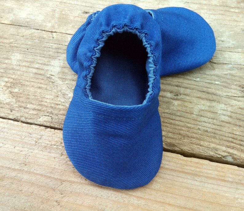 Op de afbeelding: Een paar felblauwe babyschoentjes van zachte stof. De schoenen hebben een elastische opening en een ronde neus. Ze liggen op een lichtbruin houten oppervlak.
