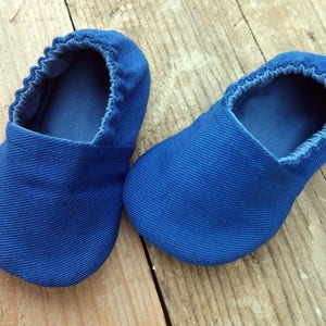 Op de afbeelding: Een paar blauwe babyschoentjes van een textielstof. De schoentjes hebben een ronde neus en elastiek rond de opening. Geplaatst op een houten oppervlak.