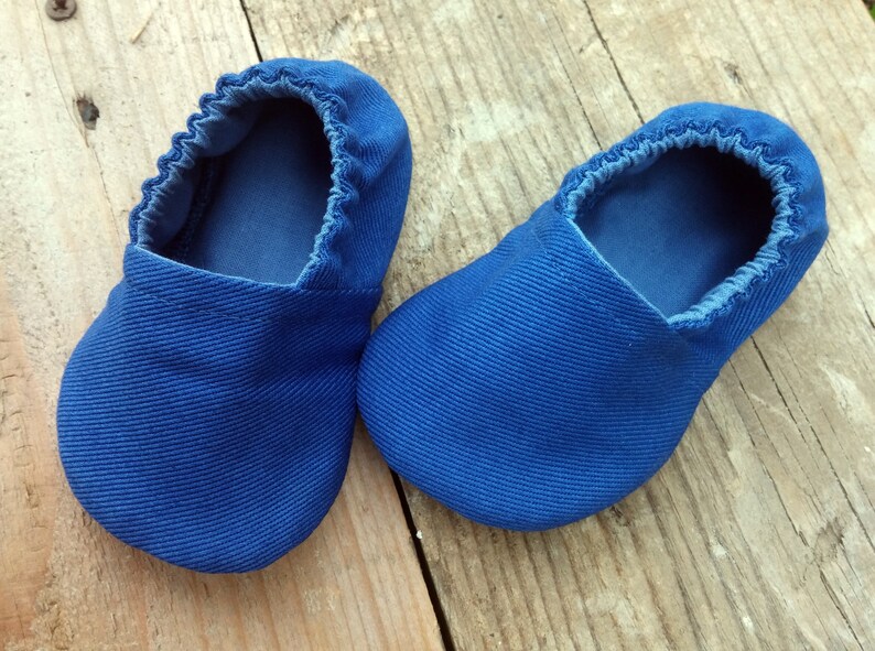 Op de afbeelding: Een paar blauwe babyschoentjes van een textielstof. De schoentjes hebben een ronde neus en elastiek rond de opening. Geplaatst op een houten oppervlak.