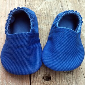Op de afbeelding: Een paar blauwe babyslofjes van een textielstof. De slofjes hebben een ronde neus en een geplooide elastische opening. De kleur is helderblauw en de slofjes worden getoond op een houten oppervlak.