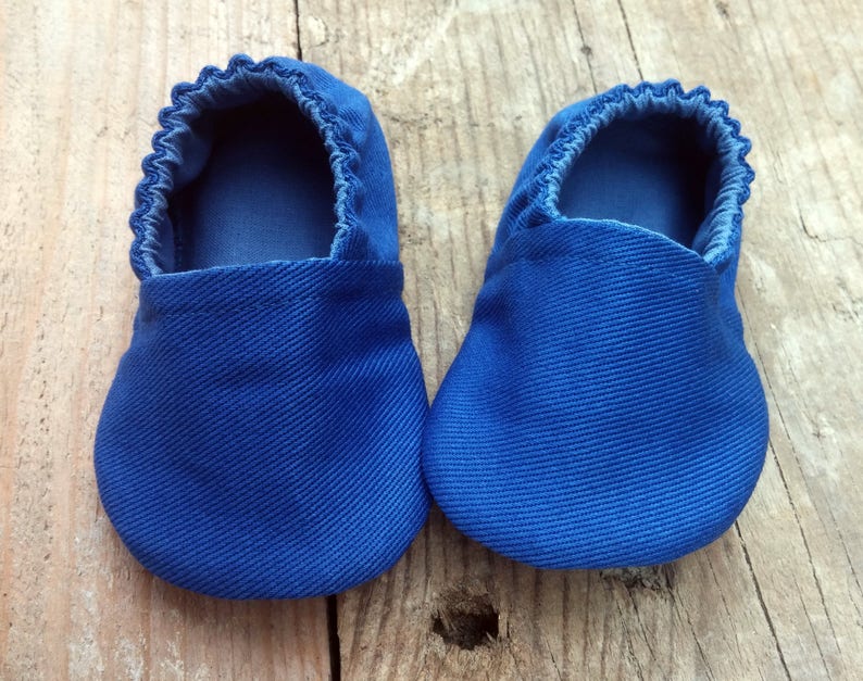 Op de afbeelding: Een paar blauwe babyslofjes van een textielstof. De slofjes hebben een ronde neus en een geplooide elastische opening. De kleur is helderblauw en de slofjes worden getoond op een houten oppervlak.