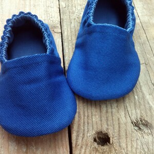 Op de afbeelding: Een paar blauwe babyschoentjes van een textielstof. De schoenen hebben een ronde neus en elastiek rond de opening. De schoenen worden op een houten oppervlak gepresenteerd.