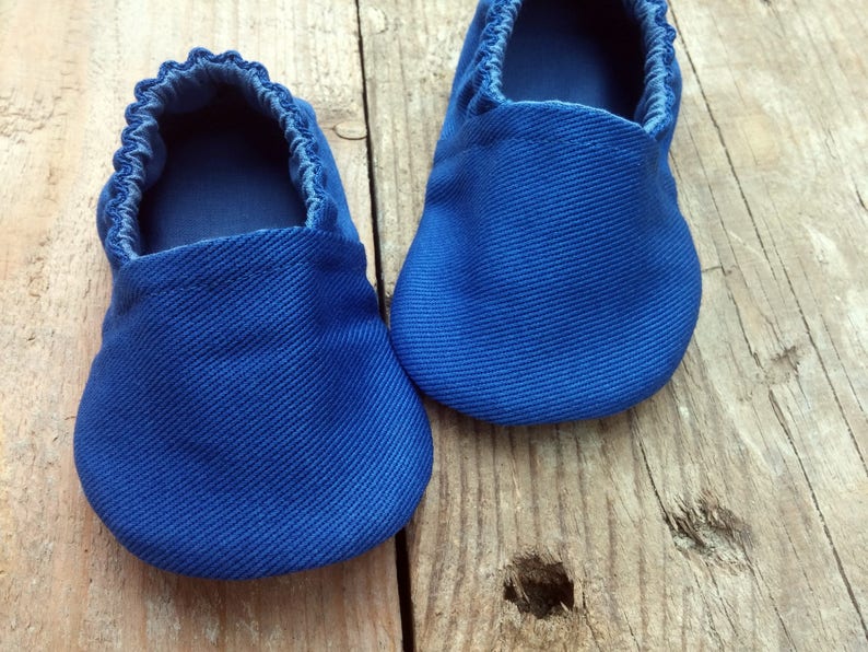 Op de afbeelding: Een paar blauwe babyschoentjes van een textielstof. De schoenen hebben een ronde neus en elastiek rond de opening. De schoenen worden op een houten oppervlak gepresenteerd.