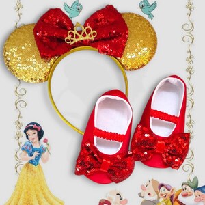 Könnte beinhalten: Rote Baby-Schuhe mit Pailletten und passenden Schleifen sowie ein goldfarbenes Stirnband mit roter Pailletten-Schleife und Ohren. Eine goldene Krone ist an der roten Schleife befestigt. Das Thema ist Schneewittchen.