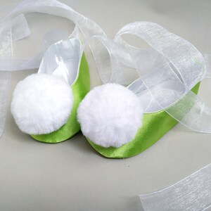 May include: A pair of lime green satin ballet slippers with white fluffy pom-poms on the toes. The slippers are tied together with a white sheer ribbon.