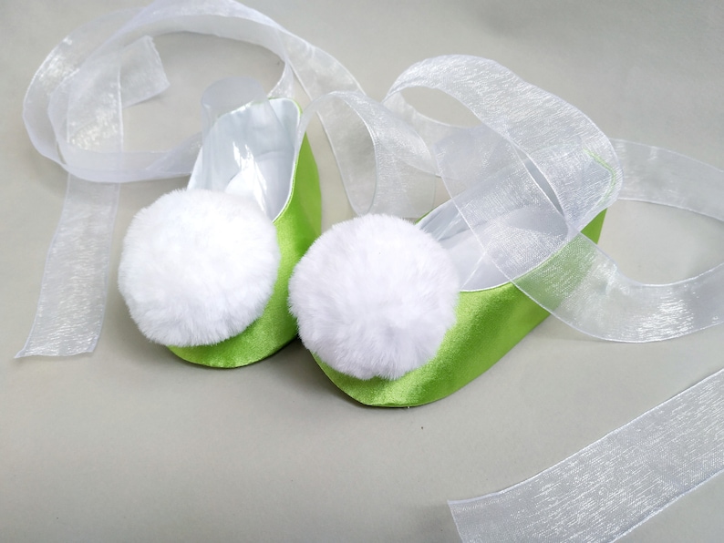 May include: A pair of lime green satin ballet slippers with white fluffy pom-poms on the toes. The slippers are tied together with a white sheer ribbon.