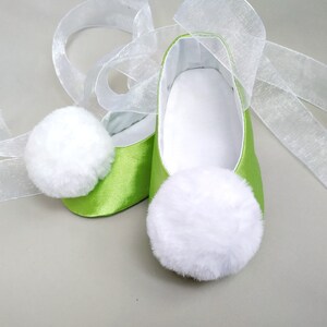 May include: A pair of lime green satin baby shoes with white fluffy pom poms on the toes. The shoes are tied with white sheer ribbon.