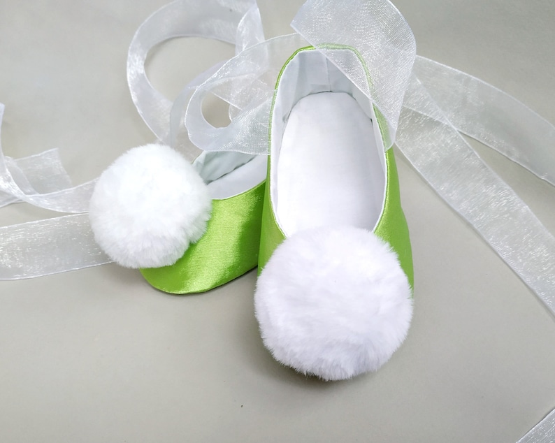 May include: A pair of lime green satin baby shoes with white fluffy pom poms on the toes. The shoes are tied with white sheer ribbon.
