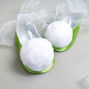 May include: A pair of green satin ballet slippers with white fluffy pom-poms and white sheer ribbon ties.