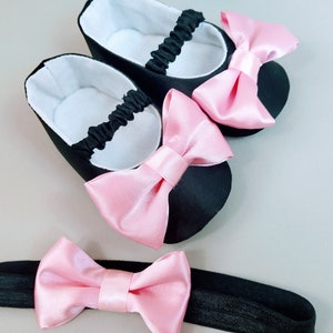 Peut inclure: Une paire de chaussures de b&eacute;b&eacute; noires avec des n&oelig;uds en satin rose et un bandeau assorti avec un n&oelig;ud en satin rose.
