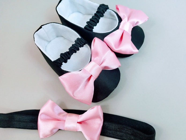Peut inclure: Une paire de chaussures de b&eacute;b&eacute; noires avec des n&oelig;uds en satin rose et un bandeau assorti avec un n&oelig;ud en satin rose.