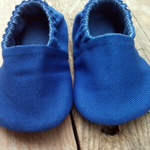 Op de afbeelding: Een paar blauwe babyschoentjes van een textielstof. De schoenen hebben een zachte, geplooide opening en een ronde neus. De schoenen worden gepresenteerd op een houten oppervlak.