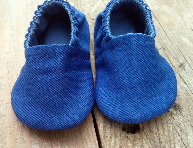 Op de afbeelding: Een paar blauwe babyschoentjes van een textielstof. De schoenen hebben een zachte, geplooide opening en een ronde neus. De schoenen worden gepresenteerd op een houten oppervlak.