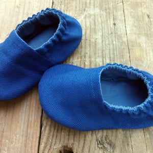 Op de afbeelding: Een paar blauwe babyslofjes van een zachte, getextureerde stof. De slofjes hebben een elastische opening en zijn ontworpen voor baby's. De schoentjes worden gepresenteerd op een rustiek houten oppervlak.