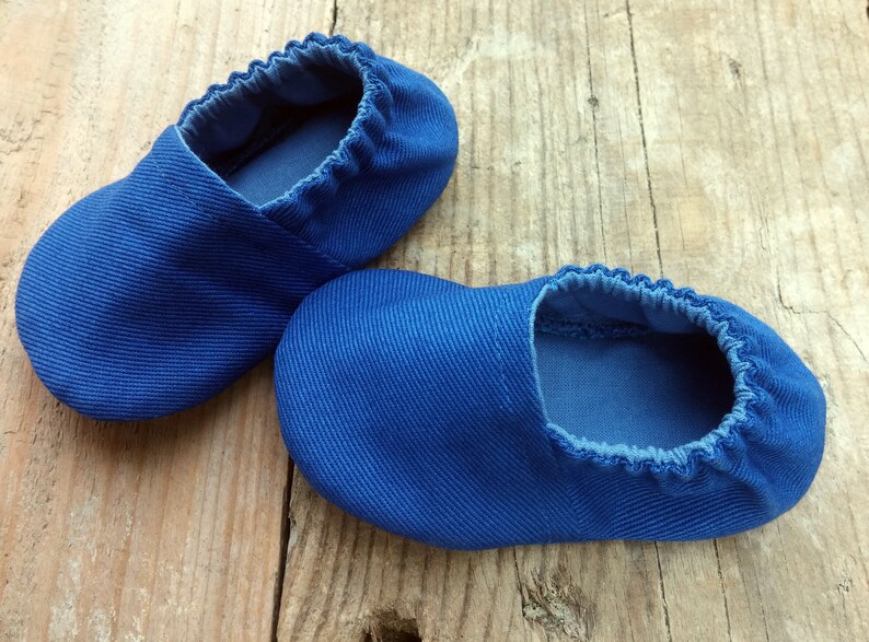 Op de afbeelding: Een paar blauwe babyslofjes van een zachte, getextureerde stof. De slofjes hebben een elastische opening en zijn ontworpen voor baby's. De schoentjes worden gepresenteerd op een rustiek houten oppervlak.