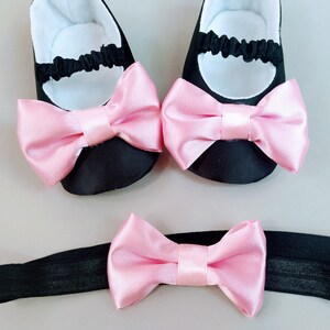 Peut inclure: Un ensemble de chaussures de b&eacute;b&eacute; noires avec des n&oelig;uds en satin rose et un bandeau noir assorti avec un n&oelig;ud en satin rose.