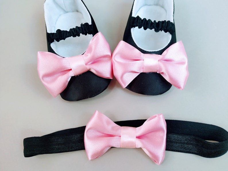 Peut inclure: Un ensemble de chaussures de b&eacute;b&eacute; noires avec des n&oelig;uds en satin rose et un bandeau noir assorti avec un n&oelig;ud en satin rose.
