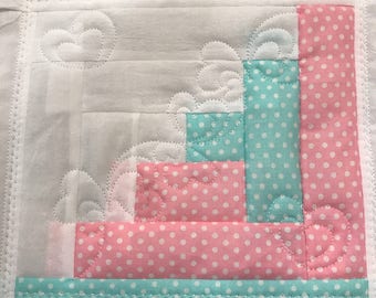May include: A square fabric quilt block featuring a log cabin pattern with alternating strips of white, light pink, and light blue polka dot fabric. The white sections are decorated with machine-stitched heart and swirl quilting patterns.