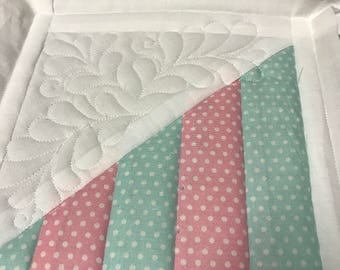 May include: A white fabric square with a quilted design. The square is made up of four smaller squares, two of which are pink with white polka dots and two of which are light blue with white polka dots. The squares are sewn together in a diagonal pattern.
