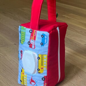 May include: A red and blue fabric wipe dispenser with a handle. The dispenser features a blue and white striped pattern with colorful illustrations of vehicles, including fire trucks, buses, and cars. A white plastic container is visible.