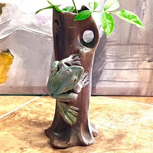 May include: A dark burgundy rose with green leaves in a brown ceramic vase. The vase has a frog sculpture on the side. The vase is on a tiled surface.