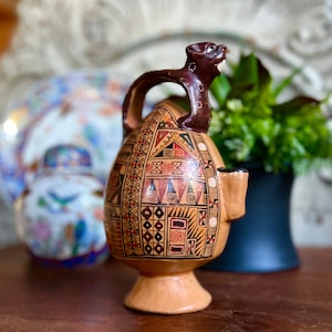 May include: A brown ceramic pitcher with a geometric pattern in red, yellow, and black. The pitcher has a handle shaped like an animal head and a small spout.