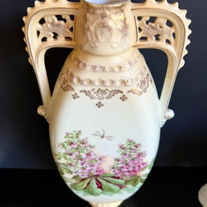 May include: A cream-colored porcelain vase with ornate gold accents and floral designs. The vase has two handles and a delicate floral pattern with pink flowers and green leaves.