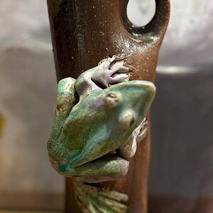 May include: A brown ceramic vase with a frog figurine. The frog is green and purple, clinging to the side of the vase. The vase has a hole and a rough texture.