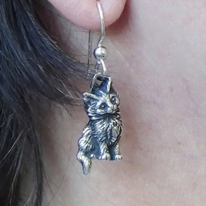 May include: A silver-toned cat-shaped earring with a detailed design. The cat is sitting upright with its tail curled. The earring hangs from a silver hook. The cat's fur and features are intricately carved.