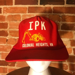 以下が含まれることがあります： 「IPK Colonial Heights, VA」という文字と、黄色いトラクターの画像が描かれた、赤と黄色のトラッカーハットです。