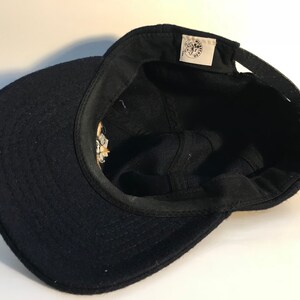 Puede incluir: Gorra de b&eacute;isbol de lana negra con un logotipo bordado en blanco y amarillo en la parte delantera. El logotipo presenta una imagen estilizada de un gato con una corona y el texto "The Cat's Meow".