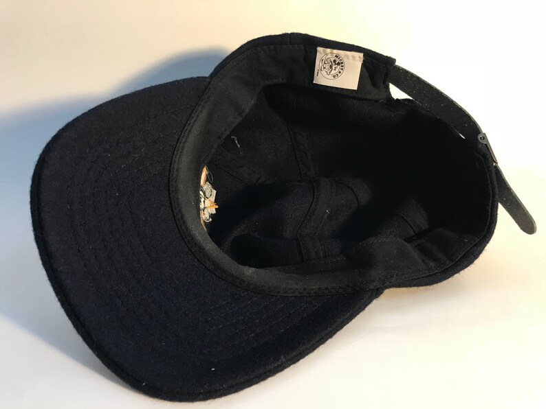 Puede incluir: Gorra de b&eacute;isbol de lana negra con un logotipo bordado en blanco y amarillo en la parte delantera. El logotipo presenta una imagen estilizada de un gato con una corona y el texto "The Cat's Meow".