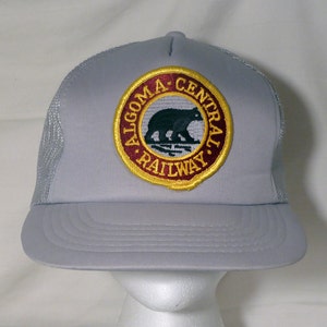 May include: A gray trucker hat with a maroon and gold embroidered patch featuring a black bear and the text "ALGOMA CENTRAL RAILWAY."