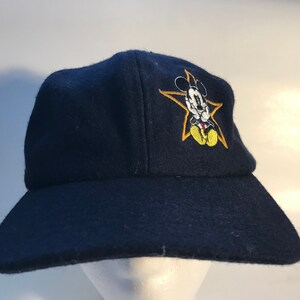 Puede incluir: Una gorra de b&eacute;isbol azul marino con una estrella amarilla y Mickey Mouse bordados en la parte delantera.