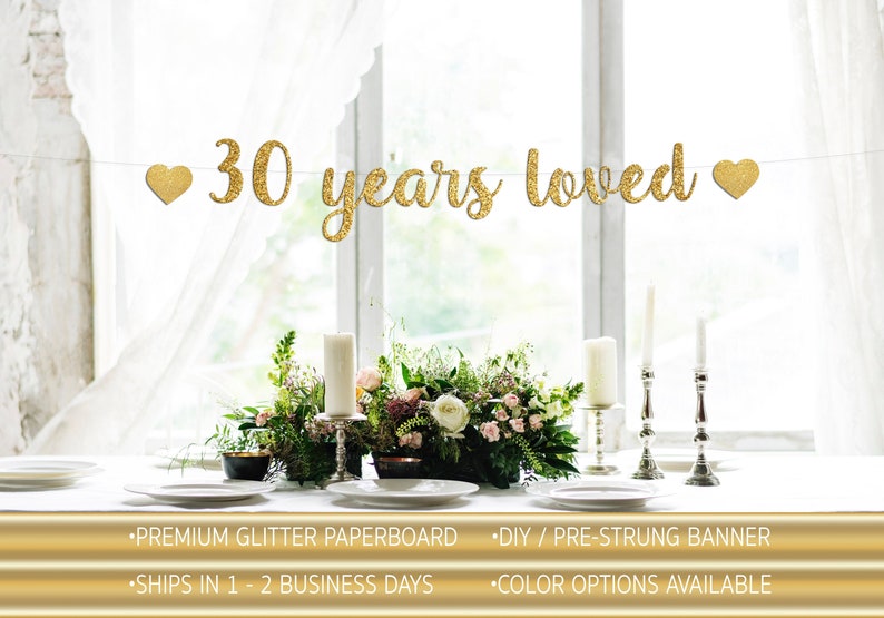 May include: Gold glitter paper banner with the text "30 years loved" and two gold glitter hearts. The banner is strung on a white string and is hanging in front of a window. The banner is on a white table with a floral centerpiece and candlesticks.