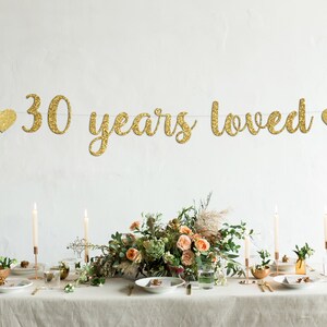 May include: Gold glitter banner with the text "30 years loved" and two gold glitter hearts on either side. The banner is hanging above a table set with candles, flowers, and plates.