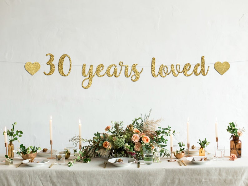 May include: Gold glitter banner with the text "30 years loved" and two gold glitter hearts on either side. The banner is hanging above a table set with candles, flowers, and plates.