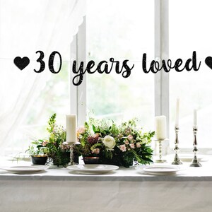 May include: Black glitter banner with white hearts and the text "30 years loved" hanging above a table setting with white tablecloth, candles, and flowers.