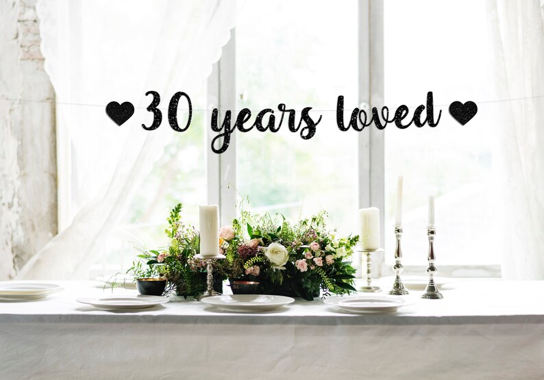 May include: Black glitter banner with white hearts and the text "30 years loved" hanging above a table setting with white tablecloth, candles, and flowers.