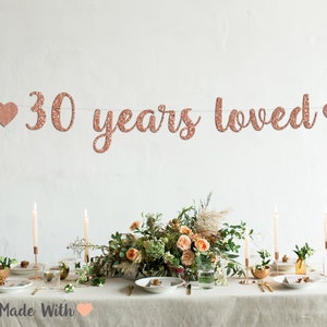 May include: Rose gold glitter banner with the text "30 years loved" and two hearts on either side. The banner is hanging above a table setting with candles, flowers, and plates.