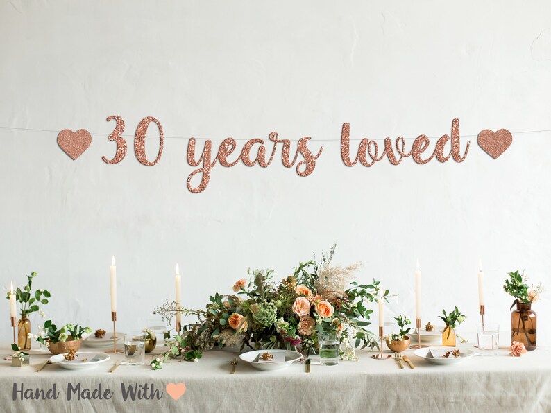 May include: Rose gold glitter banner with the text "30 years loved" and two hearts on either side. The banner is hanging above a table setting with candles, flowers, and plates.