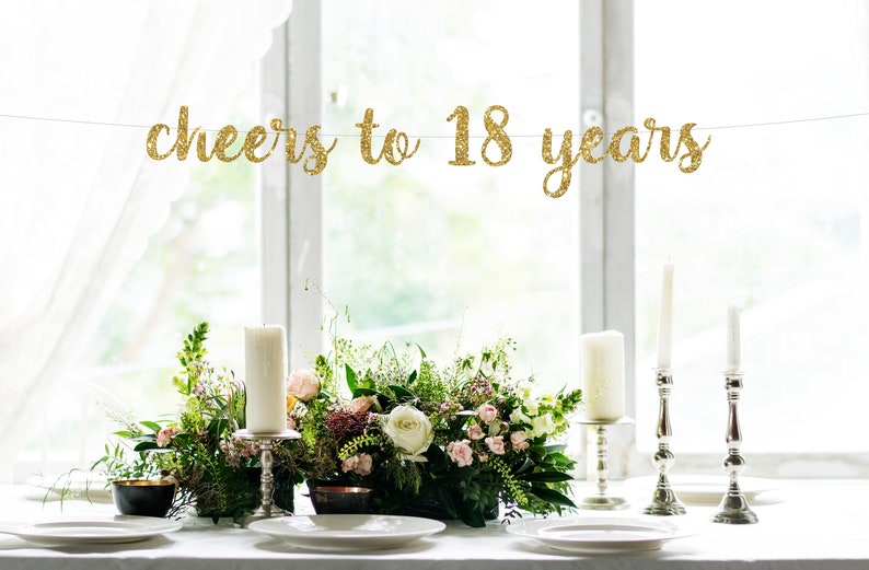 May include: A gold glitter banner that reads 'cheers to 18 years' hanging above a table setting with white candles, silver candle holders, and a floral centerpiece.